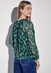 Durchsichtige, grüne Bluse mit einem abstrakten Muster, langen Ärmeln und einem gerafften Halsausschnitt, kombiniert mit blauen Jeans.