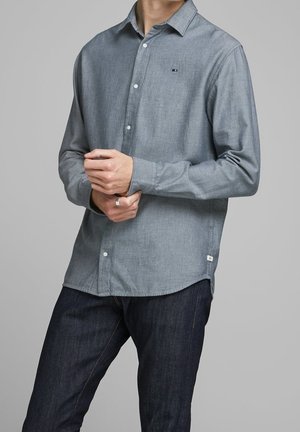 Mand iført en langærmet lysegrå skjorte med knapper og mørkeblå jeans, med hænderne foldet ved taljen.