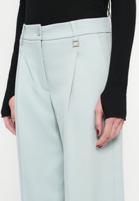 Világoskék, testhezálló nadrág, elöl hajtásokkal és egyenes szabással, téglalap alakú ezüst díszítéssel és magas derékpánttal. Fekete, hosszú ujjú felső látható.