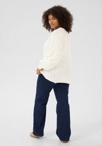 Un maglione cremoso, testurizzato con un motivo a zigzag, abbinato a jeans a zampa d'elefante blu scuro e scarpe beige, mostrato da un angolo laterale.