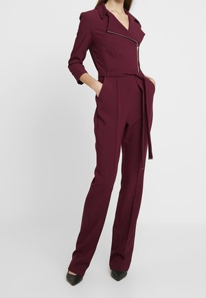 Vinrød jumpsuit med skræddersyet design, lange ærmer, bælte i taljen og sølv lynlåsdetalje. Fremstillet af glat, struktureret stof.
