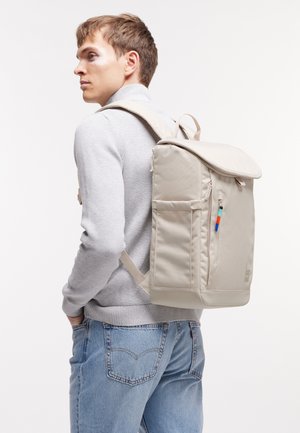 Jeune homme portant un pull gris clair et un jean bleu, portant un grand sac à dos beige avec des poches latérales et une tirette colorée.