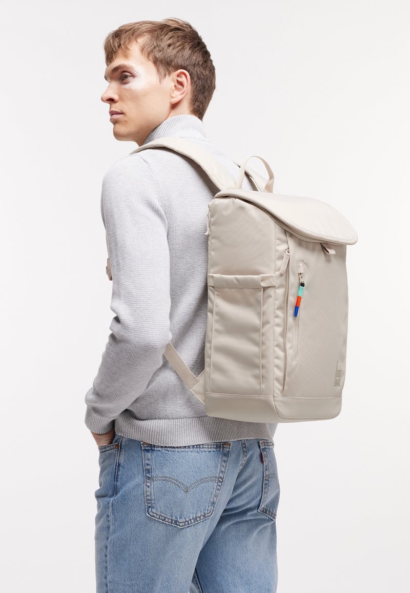 Jeune homme portant un sweat gris clair et un jean bleu, muni d'un sac à dos rectangulaire beige avec des poches latérales et une poche zippée à l'avant.