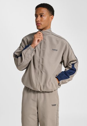 Beige Sportjacke mit hohem Kragen, marineblauen Akzenten an den Ärmeln, elastischen Bündchen und einem Frontreißverschluss. Taillierter Schnitt, kombiniert mit passenden Hosen.