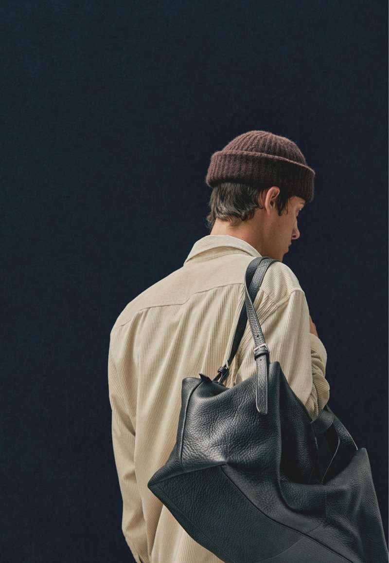 Sort lædertaske med struktureret overflade, båret på en skulder. Personen har en lys beige corduroy-skjorte og en brun strikket beanie på.