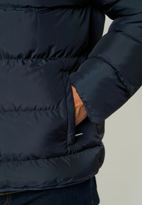 Marineblå boblejakke med horisontal søm, som har en sidelomme og en glatt tekstur. Ermene er litt puffete, noe som øker varmen.