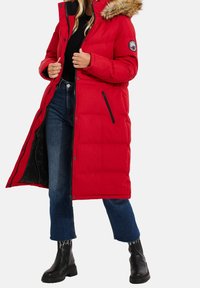 Manteau rembourré rouge avec une capuche en fourrure, poche zippée et fermetures à pression, associé à un jean foncé et des bottines noires.