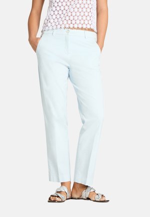 Kobieta ubrana w jasnoniebieskie spodnie o prostych nogawkach, biały krótki top z szydełkowego materiału oraz srebrne płaskie sandały z warkoczami, stojąca z rękami w kieszeniach.