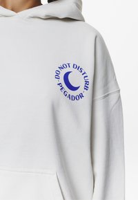 Sudadera blanca de material suave que presenta un gráfico circular azul con la frase "NO MOLESTAR PEGADOR" y un diseño de luna creciente.