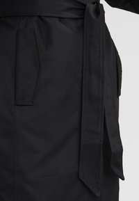 Detail černého trenčkotu s páskem na zavázání, bočními kapsami a hladkou texturou látky. Design je minimalistický s čistými liniemi.