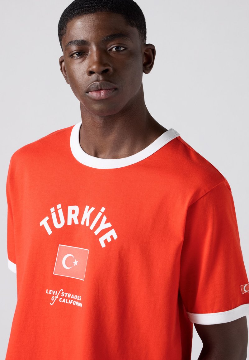Young man with short hair wearing a red T-shirt with white trim and "TÜRKİYE" text and crescent-star flag graphic on chest.