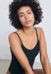 Mlada ženska kodrastih las in septum obročem, oblečena v črno majico brez rokavov, mirno gleda v kamero na enostavni ozadju.