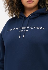 Felpa blu navy in morbido materiale con logo bianco "TOMMY HILFIGER" e testo "EST. 1985" con estremità in argento per il cordino.