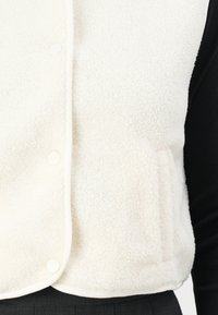 Weißer Fleece-Weste mit strukturierter Oberfläche, ausgestattet mit einer Fronttasche und Druckknopf-Verschlüssen, umrandet von einem passenden Stoffbesatz.