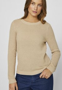 Bežani pleteni pulover s teksturiranim dizajnom, okruglim izrezom i rebrastim rukavima. Nositi preko tamnog traperica.