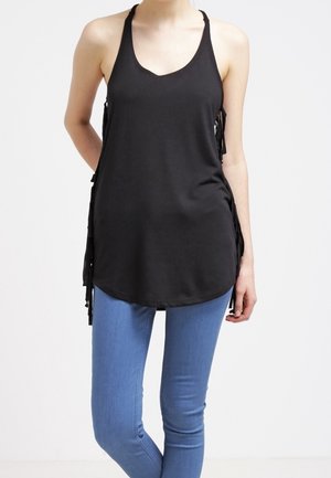 Kobieta ubrana w czarny bezrękawnik typu halter z frędzlami po bokach i jasnoniebieskie dopasowane jeansy, stojąca na tle jednolitego białego tła.