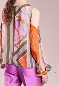 Blusa colorata con una combinazione di motivi rosa, arancione, marrone e blu, realizzata in tessuto leggero con maniche scoperte e polsini annodati.