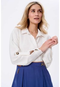 Biała bluzka z długim rękawem i podwiniętymi mankietami oraz złotymi guzikami, w połączeniu z niebieską spódnicą plisowaną z brązowym pasem.