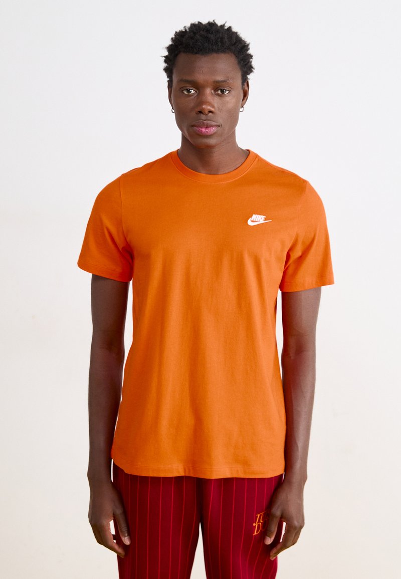 Oranžs kokvilnas t-krekls ar baltu Nike logotipu kreisajā krūtīs. Īsas piedurknes, apaļa kakla dizains, gluda tekstūra.