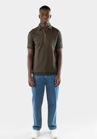 Mørkegrønn kortermet polo med glidelås i halsen, brukt sammen med lyseblå rette jeans og hvite sneakers. Glatt tekstur, avslappet passform.