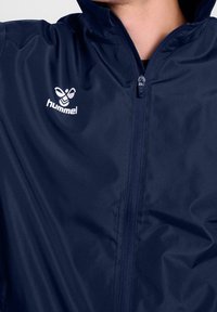 Lekka granatowa kurtka z pełnym zamkiem z przodu i wysokim kołnierzem. Na lewej stronie klatki piersiowej znajduje się biały logotyp. Gładka faktura.