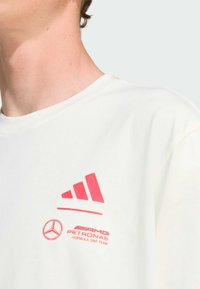 Bílý bavlněný tričko se světle růžovým logem Adidas a brandingem Mercedes-AMG Petronas na levé části hrudi.