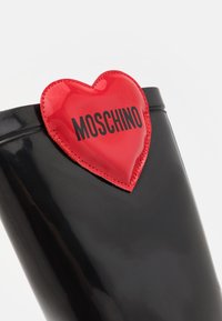 Svart blanka regstövlar med ett rött hjärtformat accent och svart text "MOSCHINO" på framsidan, som framhäver en stilren design.