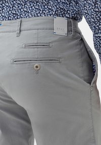 Pantaloni in cotone grigi con tasche posteriori con bottone, cuciture a contrasto e un'etichetta contrassegnata "BRAX". Tessuto testurizzato con un motivo sottile.