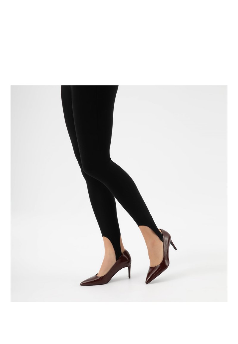 Jambes portant des leggings noirs à étrier associés à des escarpins à bout pointu marron foncé sur un fond blanc.