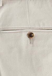 Tejido beige con una textura suave que presenta un botón decorativo en un tono más oscuro, unido con una cinta. Costura visible y un detalle de bolsillo.