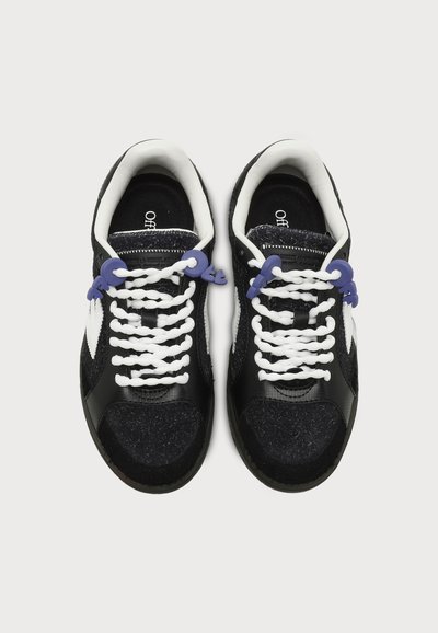 Paire de baskets noires et blanches avec des lacets blancs et des charms violets en forme de cadenas vues de dessus sur fond blanc.