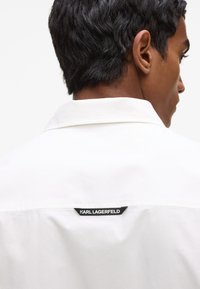 Chemise blanche avec un col classique et un patch logo noir à l'arrière avec le texte "KARL LAGERFELD". Tissu lisse avec un éclat subtil.