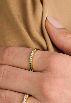 Guldbandsring med en rad av växelvis gröna och ljusblå stenar, bärs på en hand som vilar på en beige tyg.