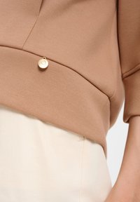 Brauner Sweatshirt mit glatter Textur, das einen abgerundeten Saum und einen kleinen runden goldenen Akzent am unteren Ende aufweist. Hellfarbener Stoff darunter.