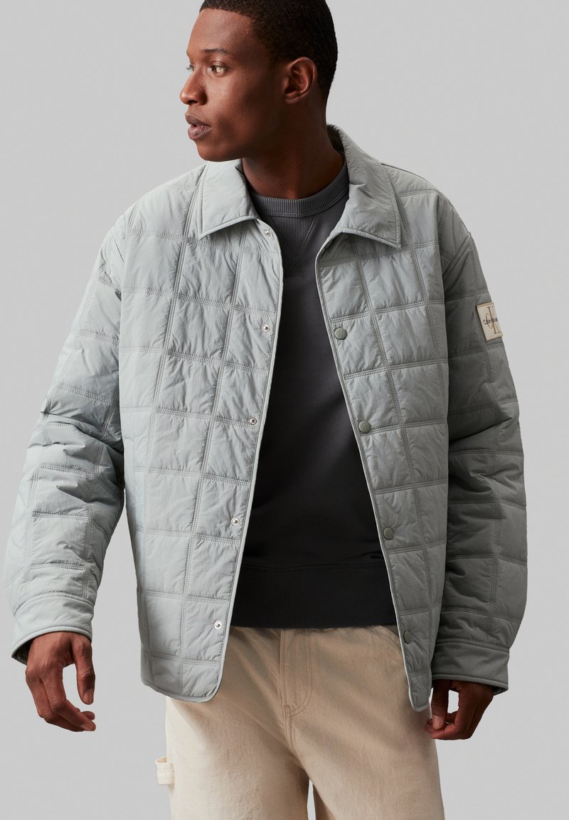 Helles grau gesteppte Jacke mit Kragen, Druckknöpfen und rechteckigem Muster. Getragen über einem dunkelgrauen Pullover und cremefarbenen Hosen.