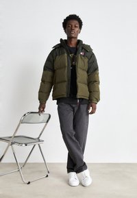 Chaqueta acolchada verde oliva con mangas más oscuras, cremallera frontal y capucha; llevada sobre un sudadera negra y combinada con jeans grises y zapatillas blancas.