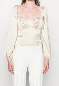Beige Satin-Top mit quadratischem Ausschnitt, gerafftem Oberteil und Puffärmeln. Die glatte Textur betont die figurbetonte Silhouette. Darunter weiße, gerippte Hosen.