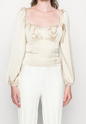 Top satin beige con scollatura quadrata, corpino arricciato e maniche a sbuffo. La texture liscia mette in risalto la silhouette aderente. Pantaloni bianchi a coste sottostanti.