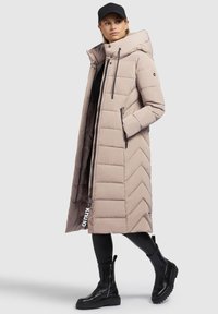 Beige gepolsterter langer Mantel mit Zickzackmuster, verstellbare Kapuze, vordere Reißverschluss-Taschen und einem Logotag. Getragen mit schwarzen Leggings und Stiefeln.