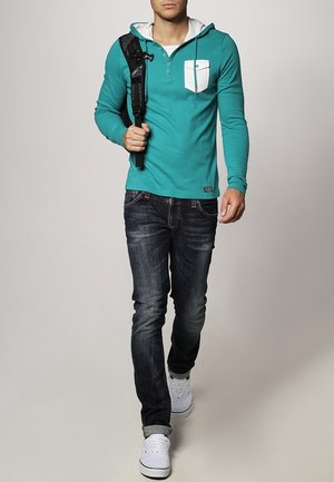 Man loopt in een teal hoodie met een witte zak, donkere jeans, witte sneakers en een zwarte schoudertas.