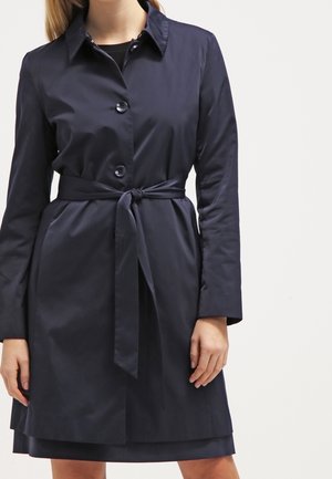 Kobieta ubrana w granatowy płaszcz trencz z paskiem, guzikami i kołnierzem, stojąca na jednolitym białym tle.