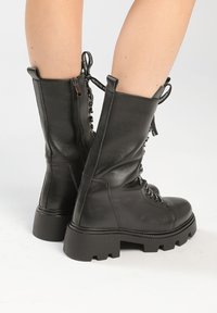 Botas de combate de cuero negras hasta media pantorrilla con suelas gruesas, cremalleras laterales y cordones negros con texto blanco, usadas sobre piernas desnudas.