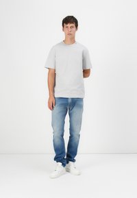 Gray short-sleeve t-shirt with a small red accent, paired with light blue jeans and white sneakers. Model standing against a white backdrop.
