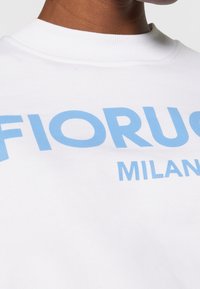Sweatshirt blanc avec un imprimé graphique bleu portant l'inscription "FIORUCI MILAN" en police audacieuse et moderne. Texture lisse et col rond.