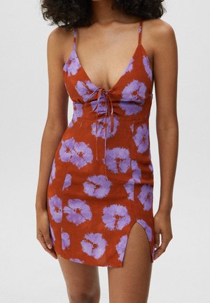 Femme portant une mini-robe sans manches orange rouille avec des motifs floraux violets et une fente à l'avant, nouée à la poitrine avec des bretelles fines.