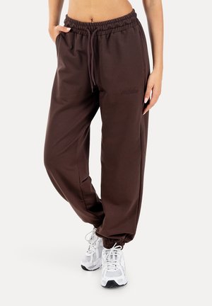 Vrouw draagt donkerbruine joggers met een trekkoord en elastische boorden, samen met witte sportschoenen, staand met handen in de zakken.