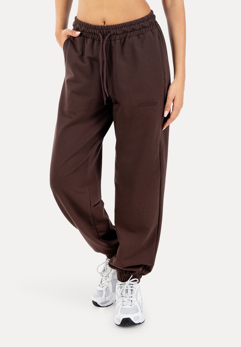 Γυναίκα που φοράει σκοτεινά καφέ joggers με κορδόνια και ελαστικές μανσέτες, καθώς και λευκά αθλητικά παπούτσια, στέκεται με τα χέρια στις τσέπες.