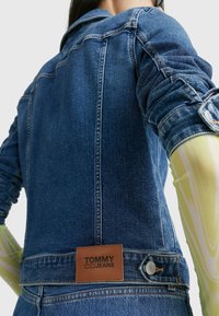 Denim jacket featuring a structured design, blue color, subtle fading, and a brown leather tag with "TOMMY JEANS" at the back waistband.