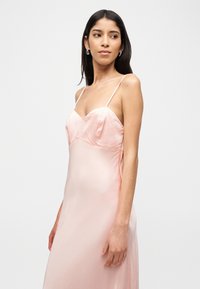 Pikkade tumedate juustega naine, kes kannab õrna roosa satiinist õhukeste rihmadega kleiti, vaadates veidi kõrvale ühevärvilise tausta ees.