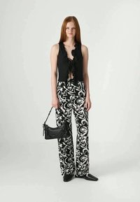 Mulher com cabelo longo e vermelho, vestindo uma blusa preta sem mangas com babados, calças largas com padrão em preto e branco, sapatos pretos de bico fino, segurando uma bolsa preta.